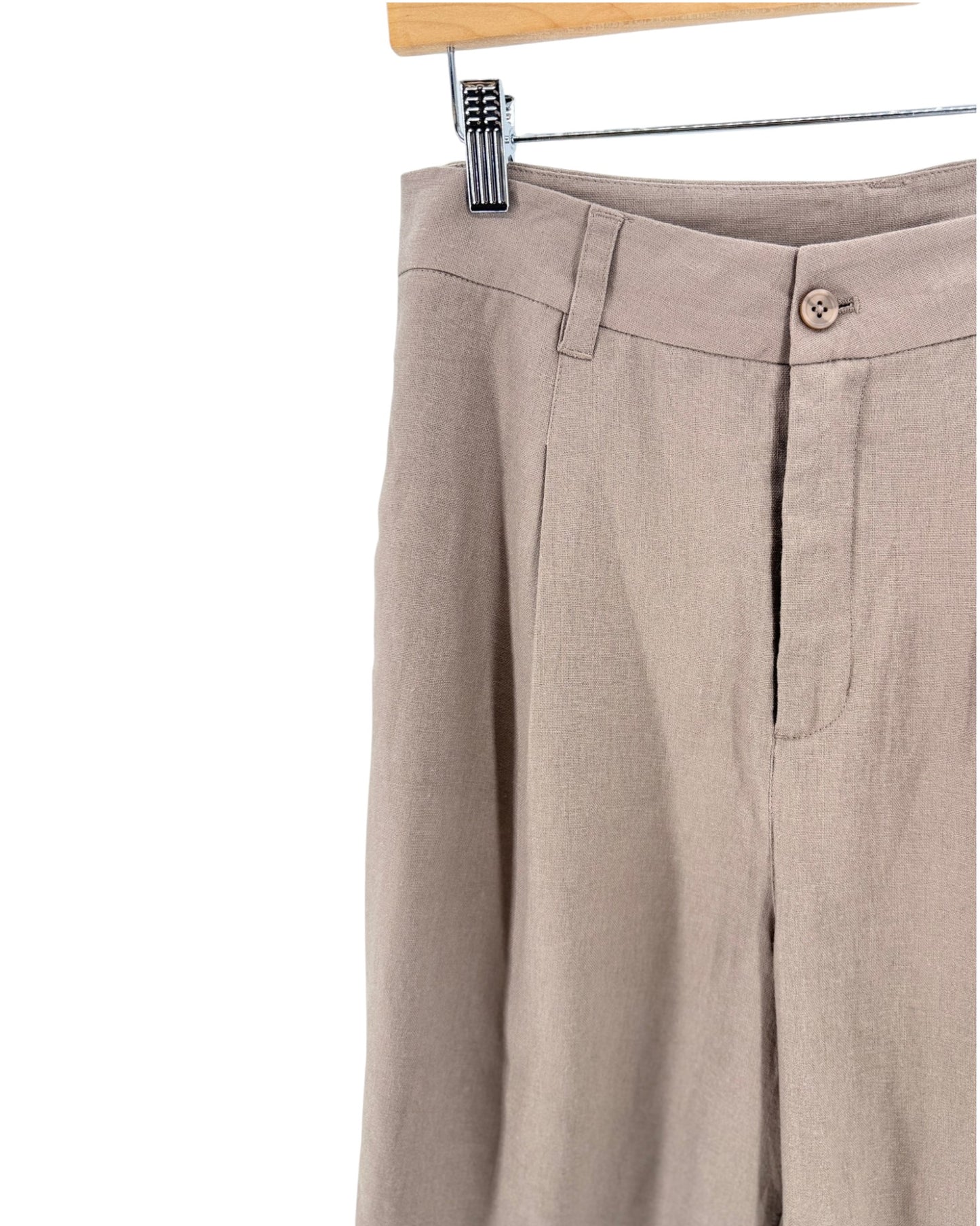 Close-up of beige linen shorts hanging from a wooden hanger with a metal clip, showing the waistband, front fly, and side pocket on a white background.