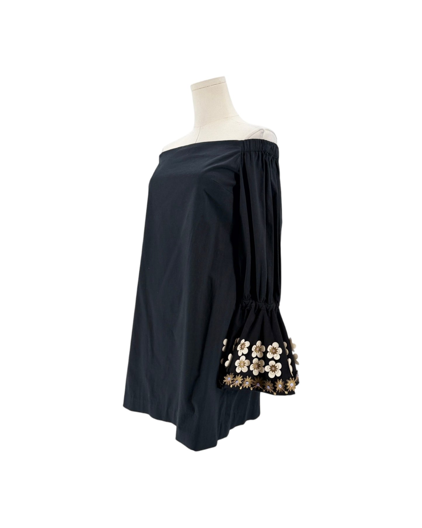 A black off-the-shoulder blouse displayed on a mannequin torso, featuring dramatic bell sleeves with a decorative floral applique trim at the cuffs.