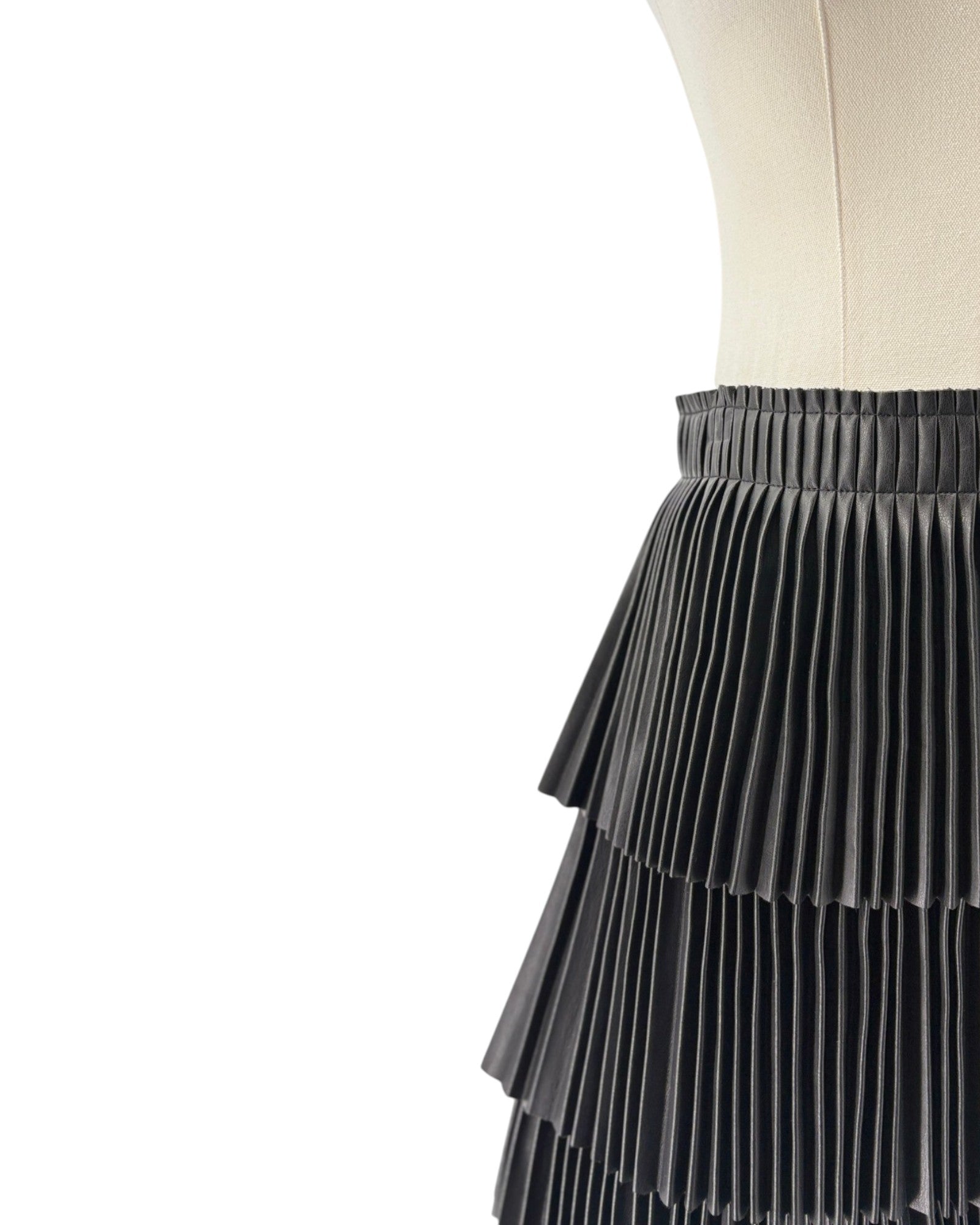 A close-up of a black pleated skirt displayed on a cream dress form against a white background.