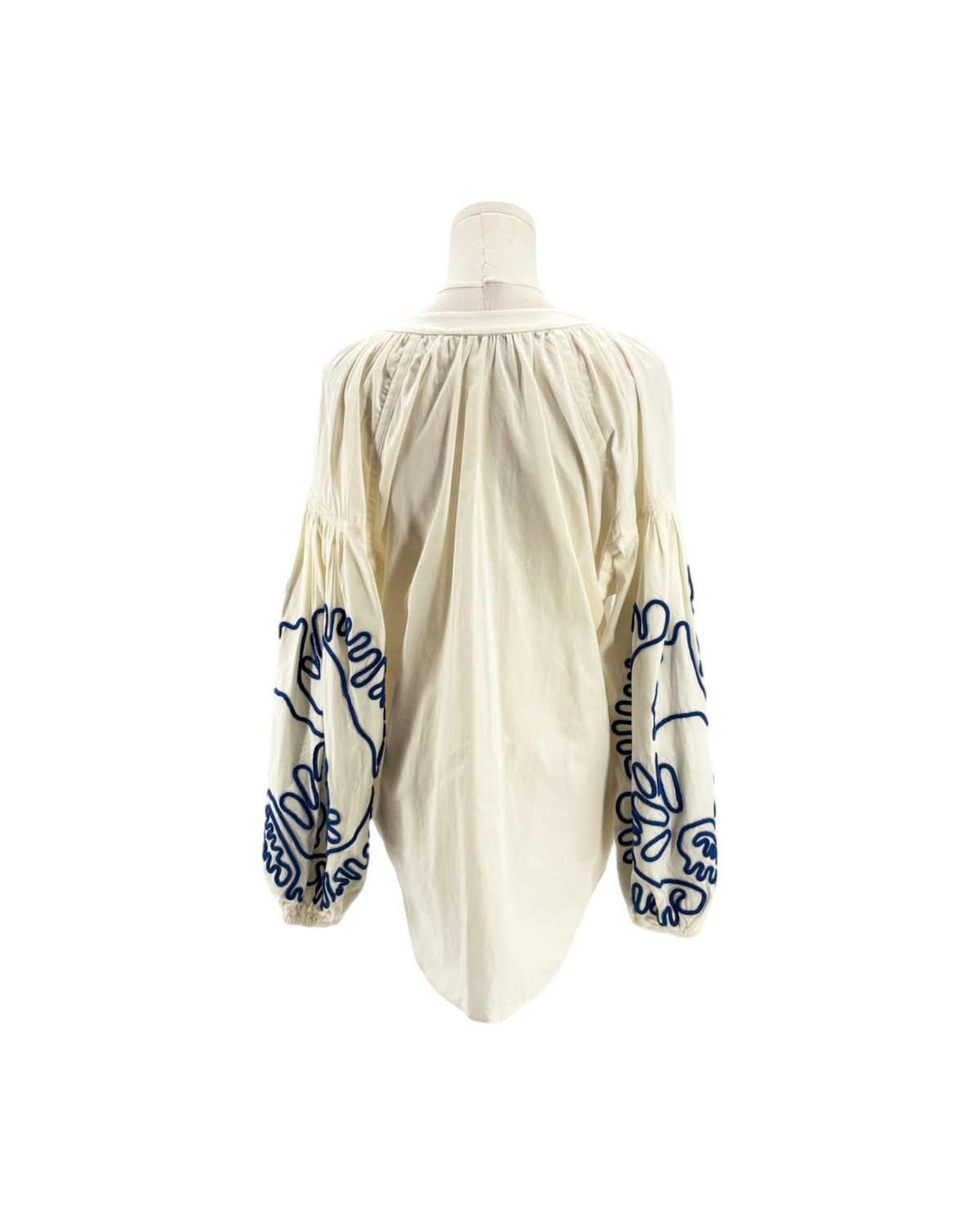 An off-white or cream-colored blouse with dramatic puffed sleeves, featuring dark blue embroidered floral and script patterns on the sleeves, displayed on a mannequin against a white background.