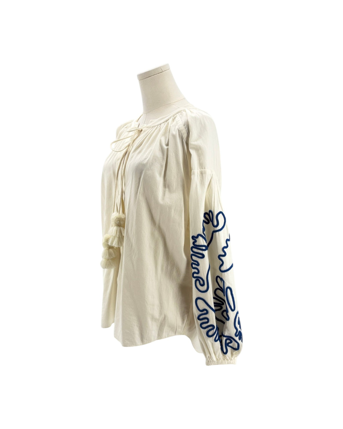 An off-white or cream-colored blouse with dramatic puffed sleeves, featuring dark blue embroidered floral and script patterns on the sleeves, displayed on a mannequin against a white background.