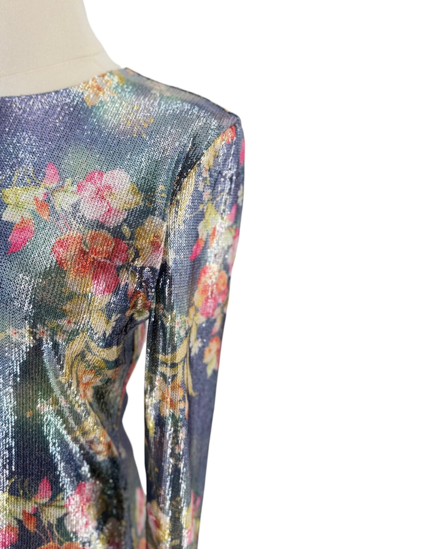 A close-up of a long-sleeve top on a mannequin, featuring a shimmering, metallic floral pattern with pink, yellow, and green tones over a dark blue-gray base.