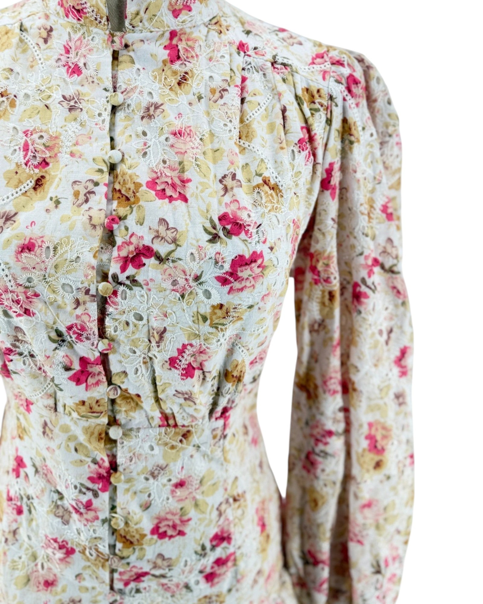 A close-up of a long-sleeve dress on a mannequin, featuring a vintage floral pattern in pink, red, and yellow on a cream background with lace applique and a button-front closure.