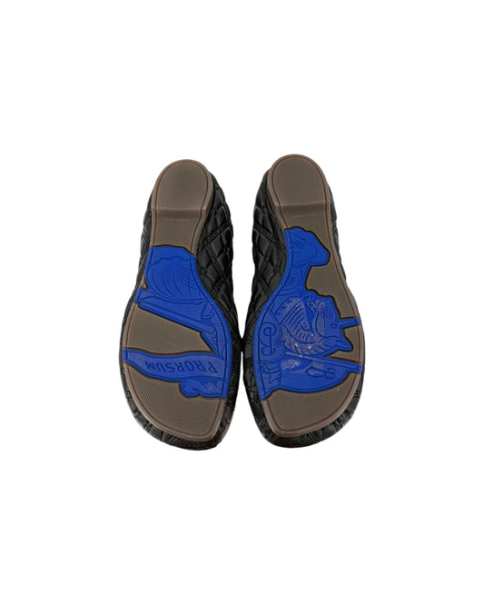 Top-down view of a pair of black quilted slip-on shoes with brown soles featuring a prominent blue embossed Greek key pattern and the word 'MASHURA'.