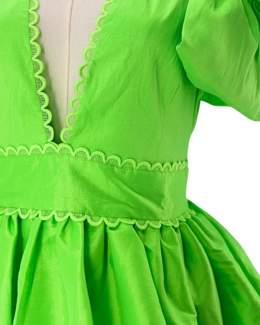 Close-up of a bright lime green dress on a mannequin, showing the deep V-neckline with matching scalloped trim, a gathered waistband, and a pleated skirt against a white background.