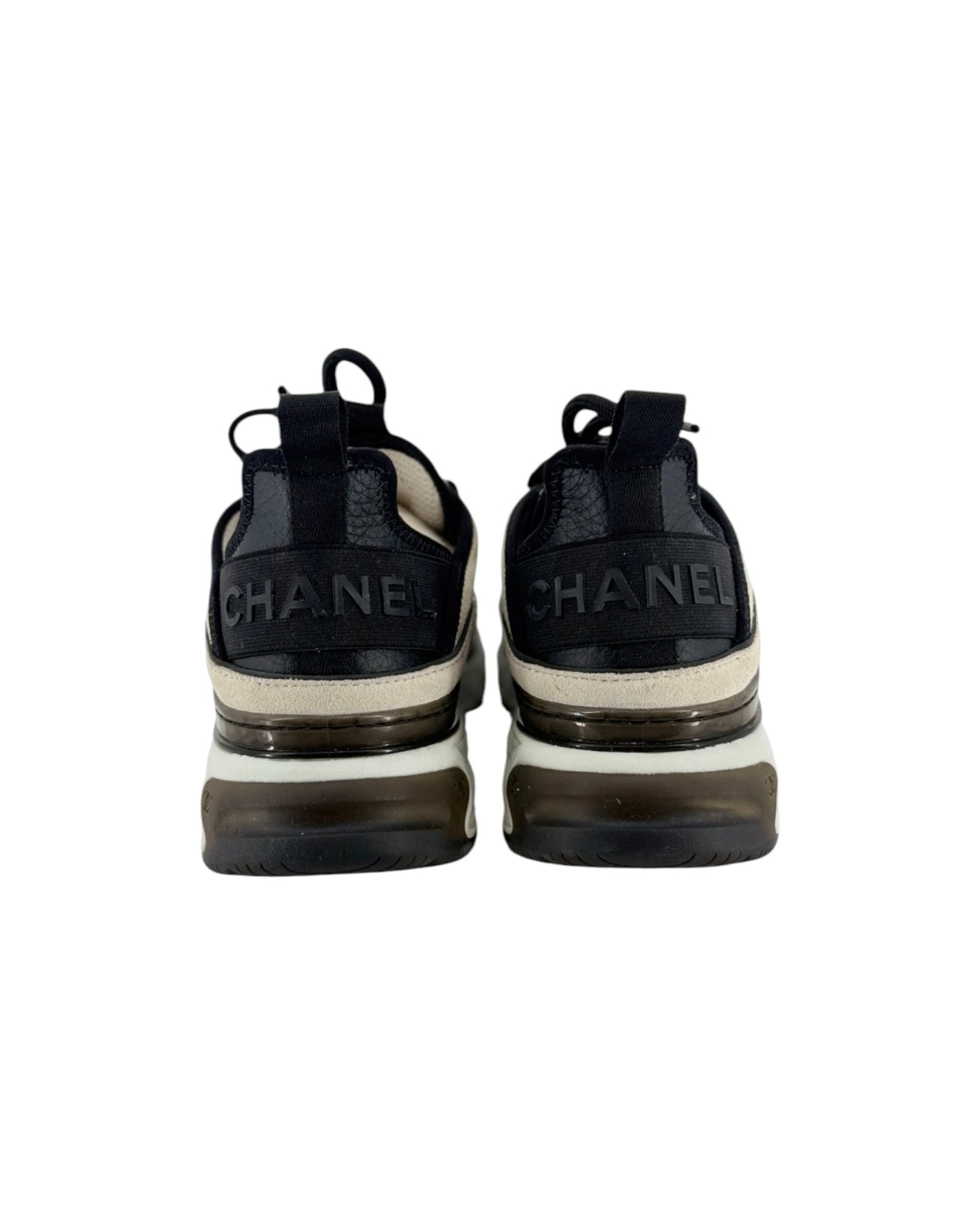 A pair of Chanel low-top sneakers shown from the back, featuring a black leather upper with 'CHANEL' lettering on the heel, a cream midsole stripe, and a chunky translucent brown and black sole.