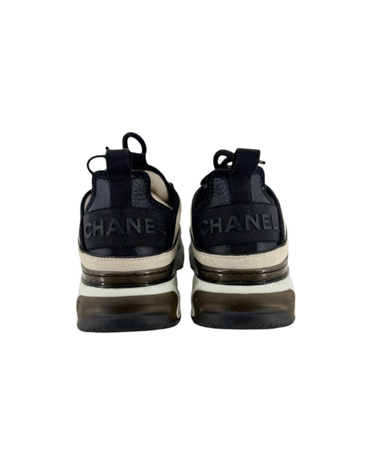 A pair of Chanel low-top sneakers shown from the back, featuring a black leather upper with 'CHANEL' lettering on the heel, a cream midsole stripe, and a chunky translucent brown and black sole.