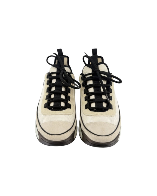 A pair of chunky low-top sneakers in off-white and beige suede and mesh, featuring prominent black laces and black soles, shown from a front-facing perspective on a white background.