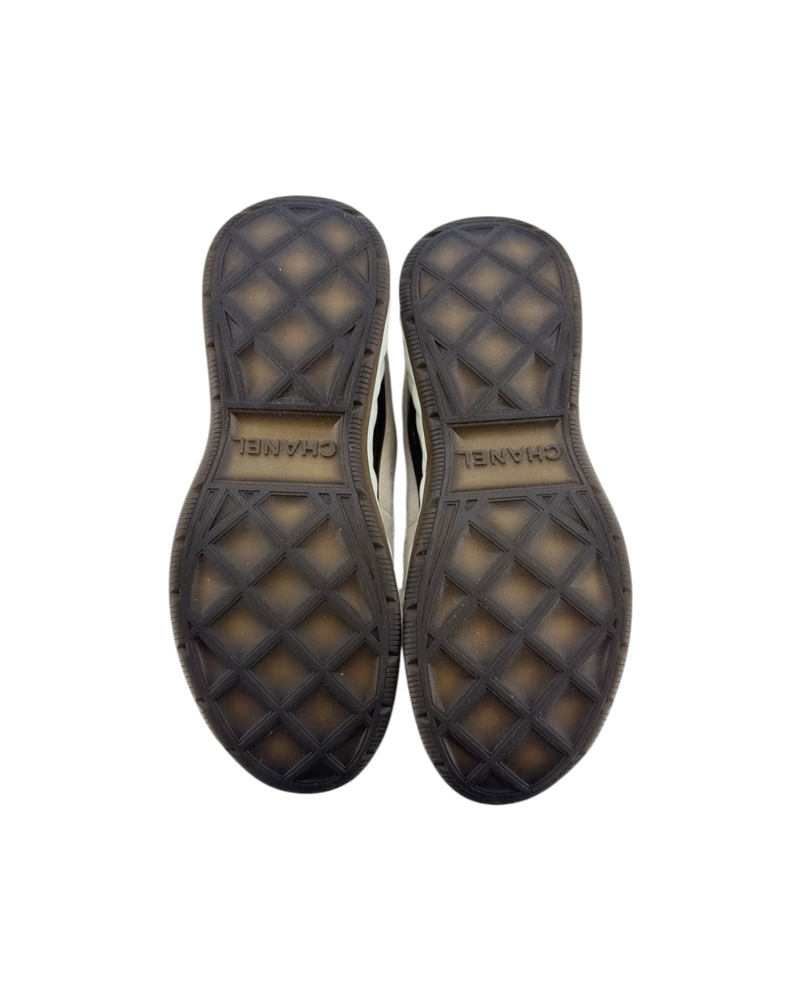 Top-down view of a pair of shoes showing the textured black rubber soles with a diamond pattern and the brand name Chanel printed on the center.