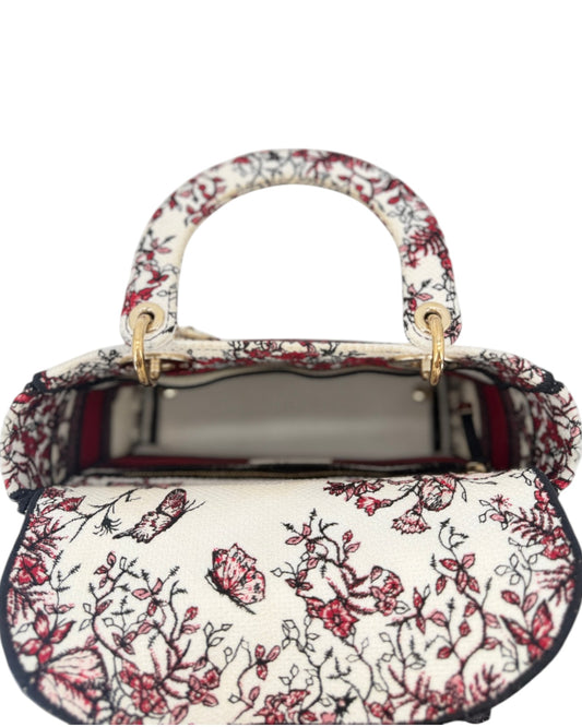 A structured handbag with a cream-colored base and an intricate red and black floral embroidery pattern, featuring gold-tone hardware and top handles.