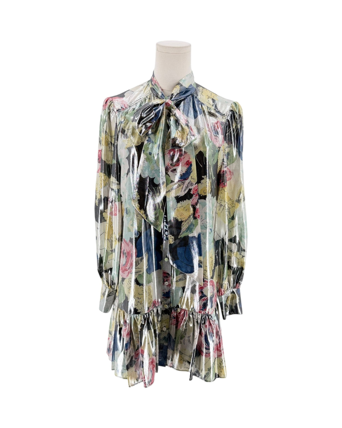 A long-sleeve, pleated blouse with a tie-neck bow, displayed on a mannequin. The shirt features a shiny, metallic finish and a multicolor floral print with green, pink, blue, and yellow tones on a light base.