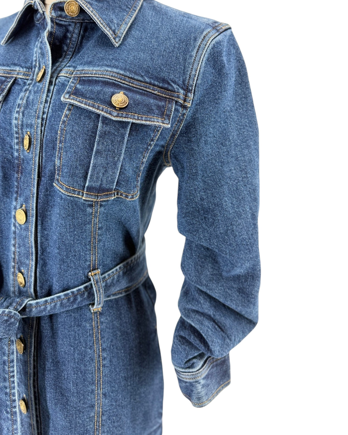 Close-up of a blue denim shirt dress with gold-tone buttons, a pointed collar, chest flap pockets, and a matching waist tie belt, shown on a white background.