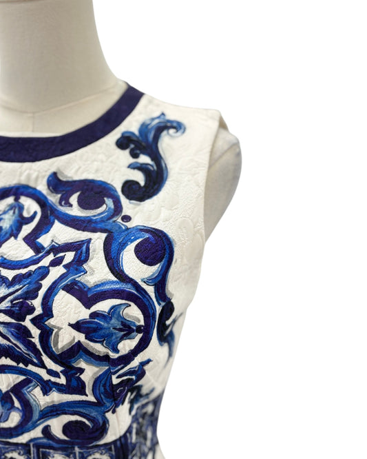 Close-up of a sleeveless white dress or top on a mannequin, featuring an ornate blue embroidered scroll pattern and a dark blue crew neckline.