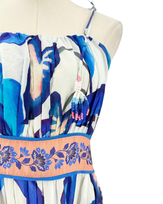 A close-up of a sleeveless dress displayed on a mannequin torso against a white background. The dress has an abstract blue, navy, and white print with a peach-colored waistband featuring a blue floral border and a beaded tassel detail.