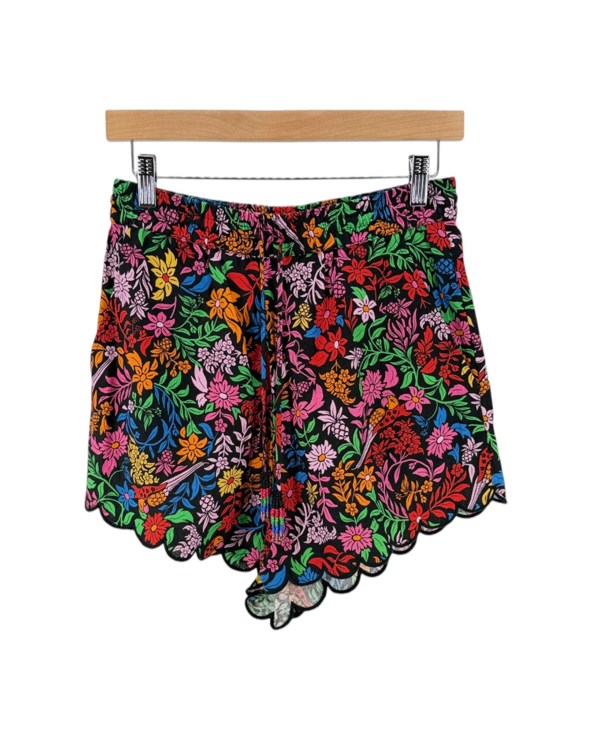 A pair of women's shorts featuring a vibrant, multicolored floral print with a scalloped hem, hanging from a wooden hanger with metal clips against a plain white background.