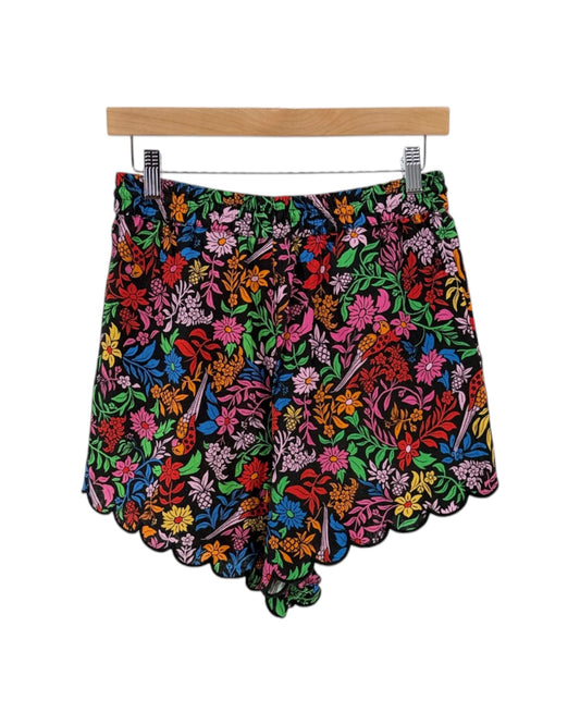 A pair of women's shorts featuring a vibrant, multicolored floral print with a scalloped hem, hanging from a wooden hanger with metal clips against a plain white background.