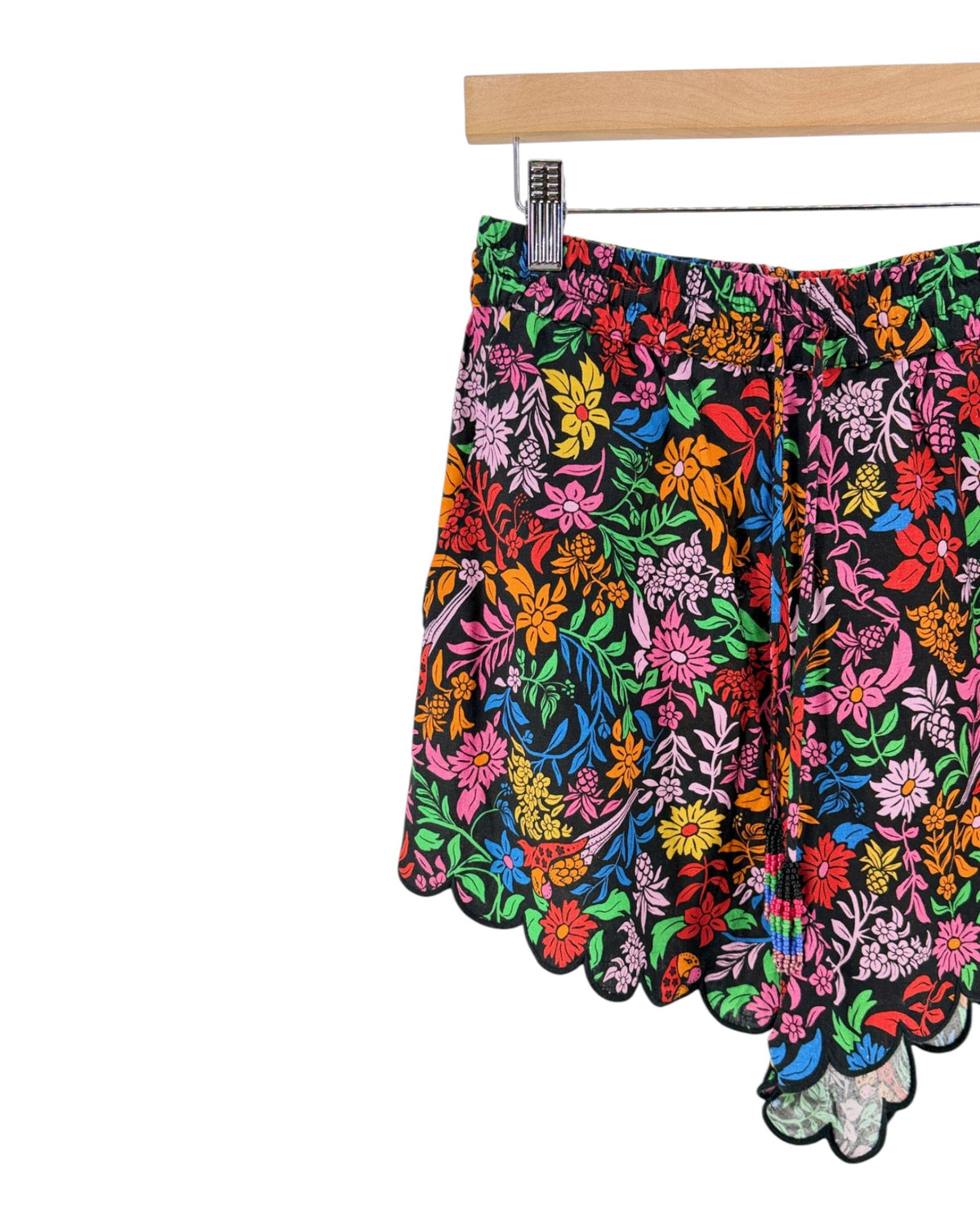 A pair of women's shorts featuring a vibrant, multicolored floral print with a scalloped hem, hanging from a wooden hanger with metal clips against a plain white background.