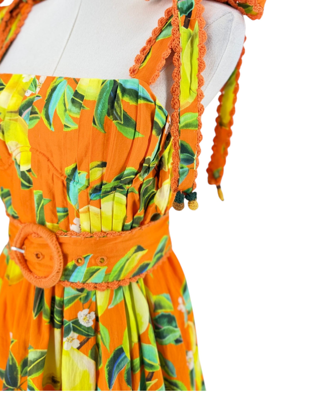 An orange sleeveless dress with a yellow and green lemon print, featuring tie-shoulder straps, a smocked bodice, and a matching orange belt with a round buckle, displayed on a white mannequin torso against a white background.