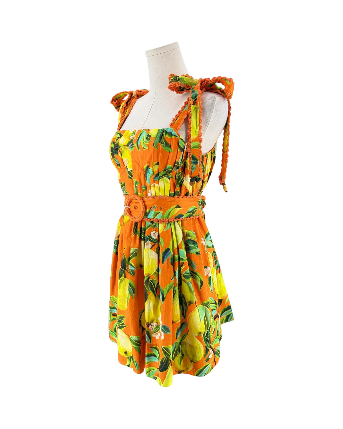 An orange sleeveless dress with a yellow and green lemon print, featuring tie-shoulder straps, a smocked bodice, and a matching orange belt with a round buckle, displayed on a white mannequin torso against a white background.