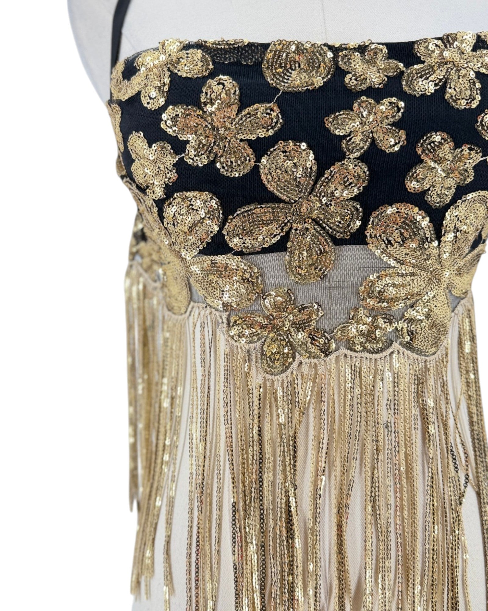 Close-up of a black and gold embellished garment on a mannequin, featuring large sequined floral motifs and long dangling fringe.
