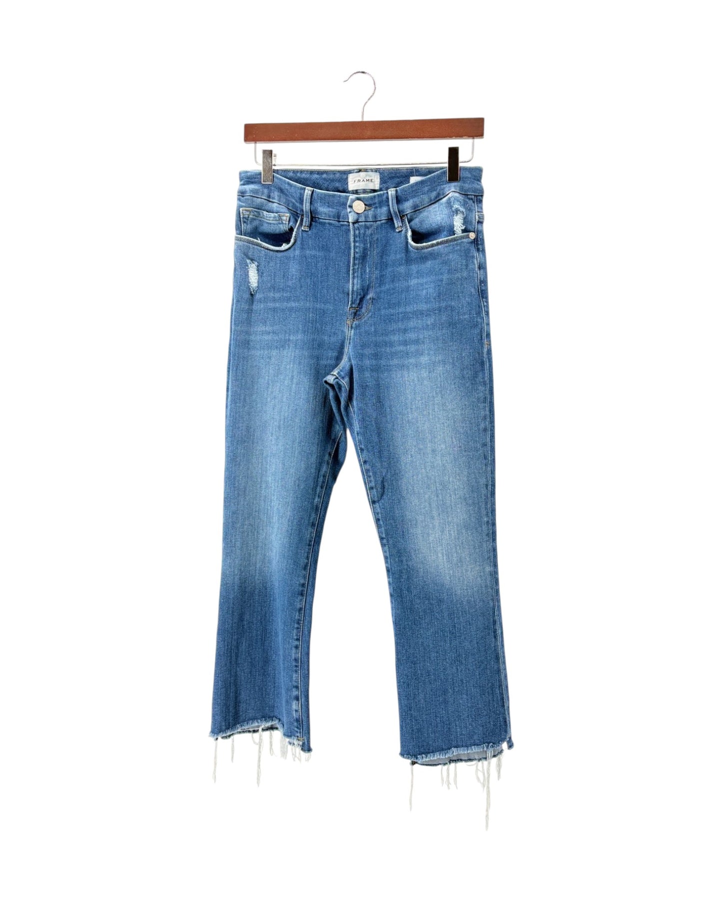 Medium wash blue denim cropped jeans with a distressed front thigh and raw frayed hems, hanging on a wooden hanger against a white background.