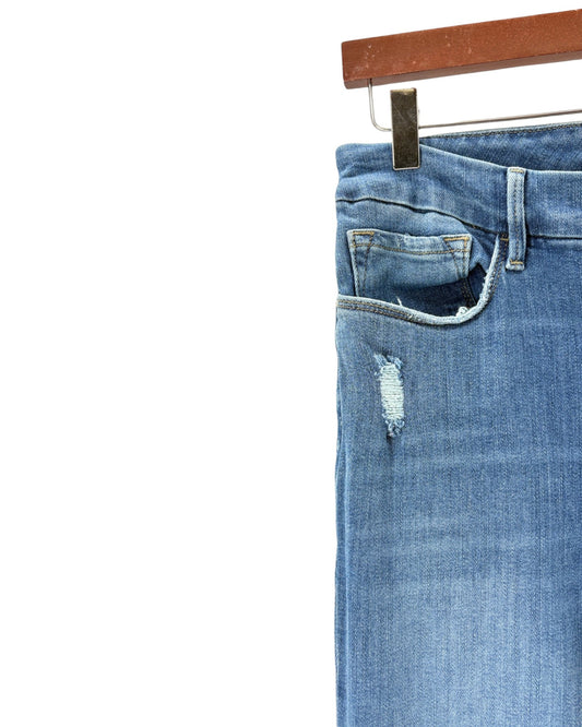 Close-up of a pair of blue denim jeans hanging from a wooden hanger, showing the waistband, belt loops, front pocket, and a distressed rip on the thigh against a white background.