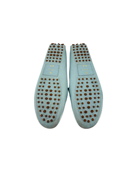 A pair of light blue leather flat shoes shown from the sole, featuring a grid of small brown studs and a small gold-tone rectangular brand plaque on each sole.