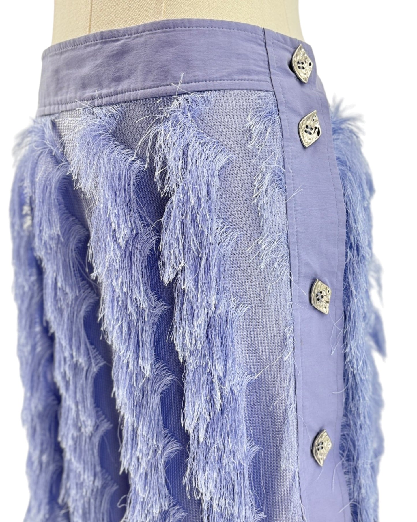 A close-up of a lavender purple skirt on a mannequin, featuring textured fringe, mesh panels, and decorative metallic buttons down the side.