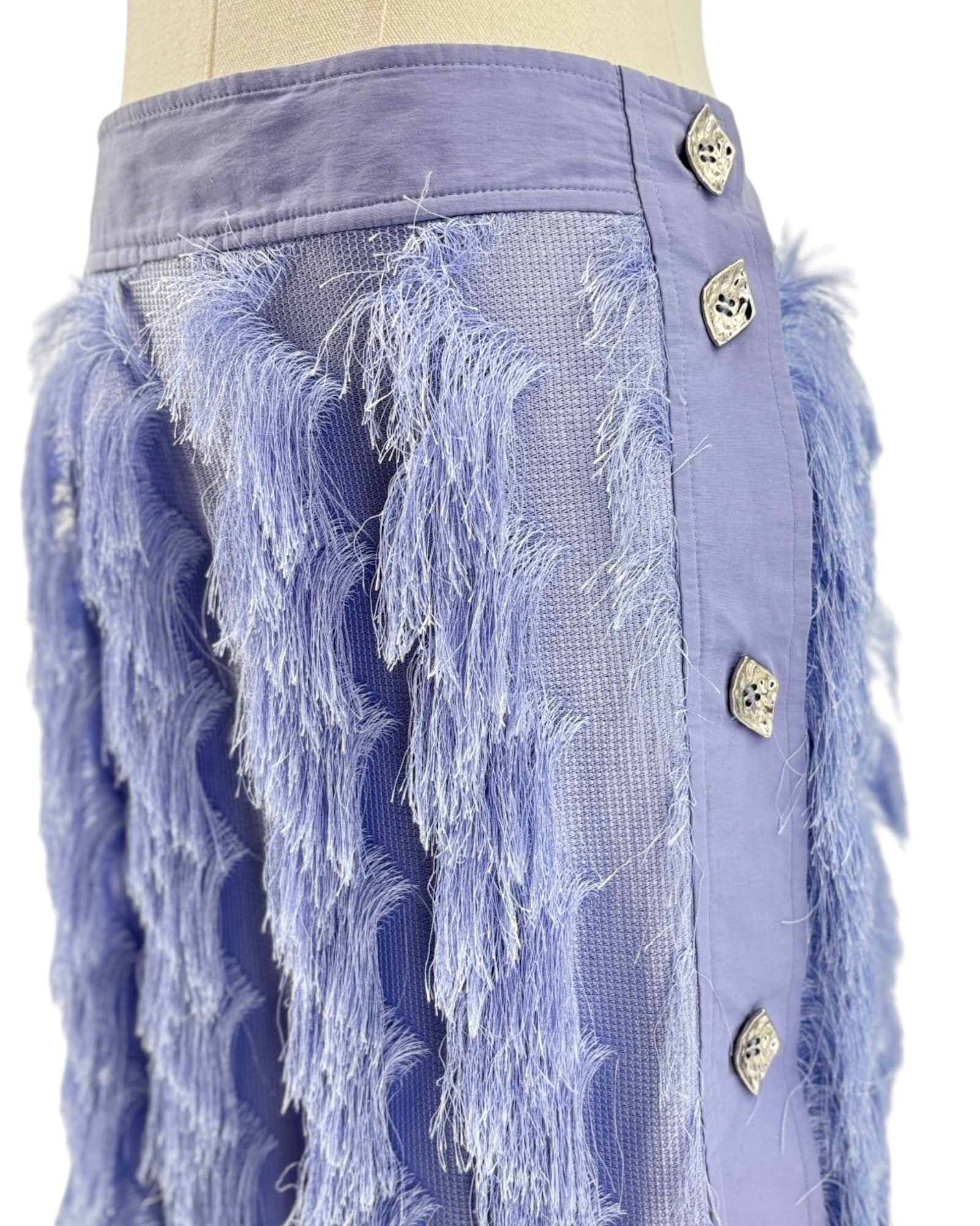 A close-up of a lavender purple skirt on a mannequin, featuring textured fringe, mesh panels, and decorative metallic buttons down the side.