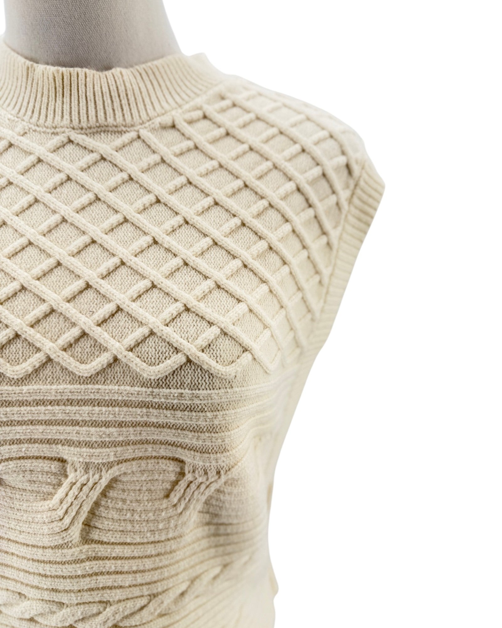Close-up of a cream-colored knit sweater vest with a textured diamond lattice pattern on the chest, displayed on a mannequin torso against a white background.