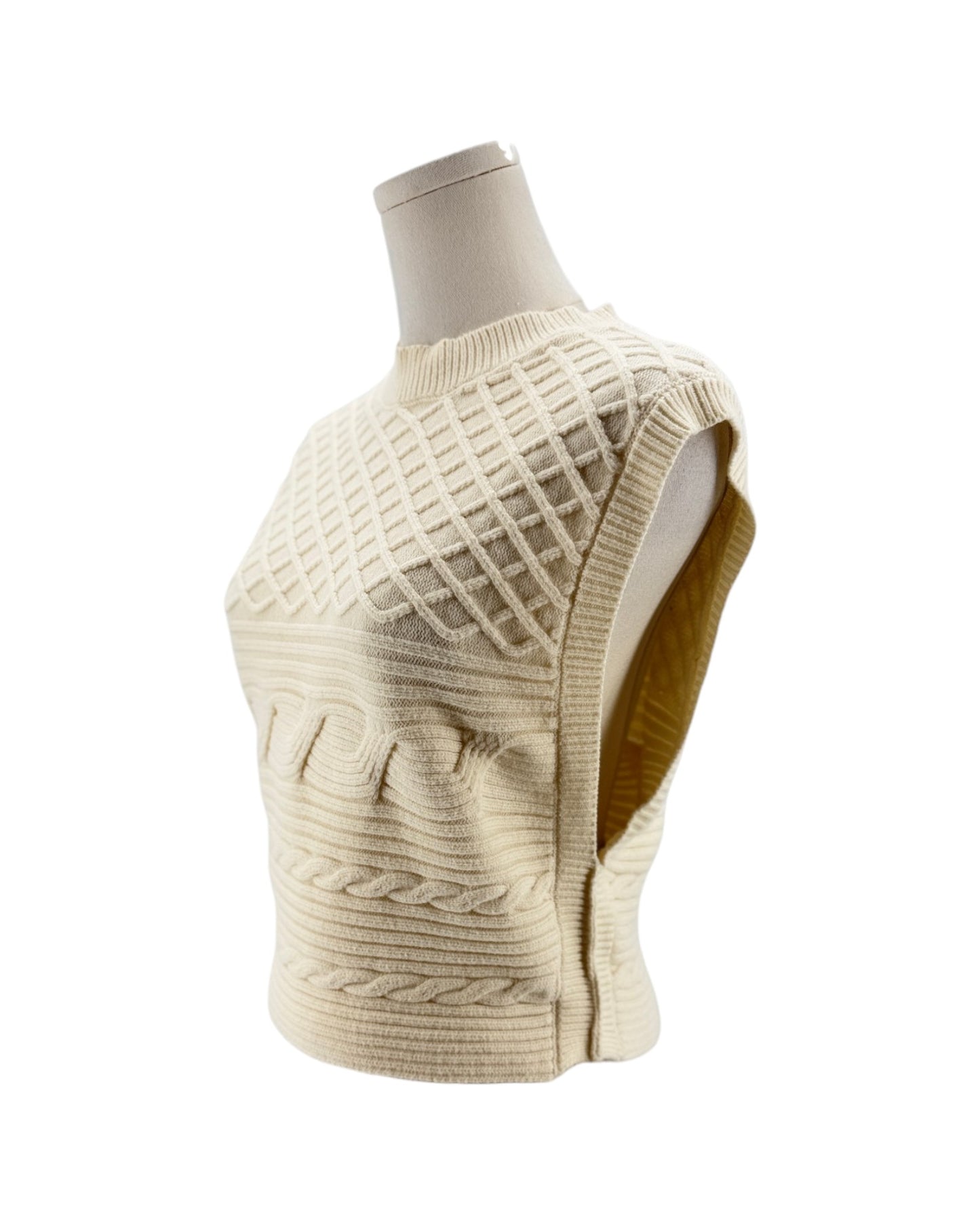 A cream-colored sleeveless knit vest with textured diamond and cable patterns, displayed on a mannequin torso against a white background.