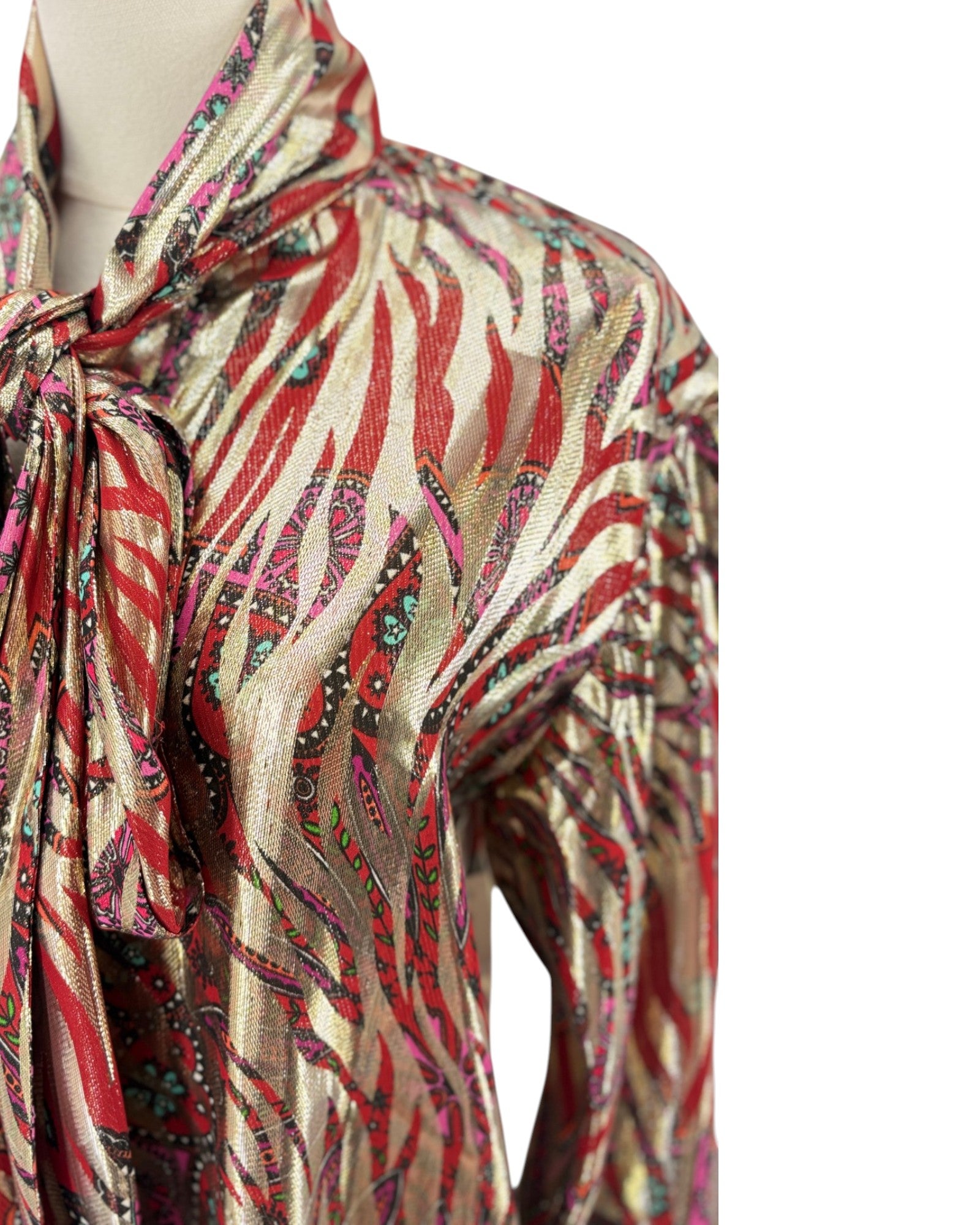 A close-up of a metallic gold and red patterned blouse on a mannequin, featuring a tie-neck detail and a paisley-like abstract print.