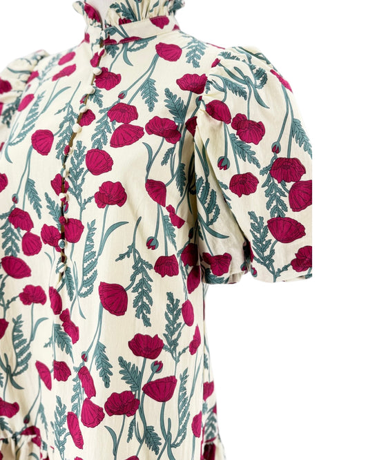 A close-up of a cream-colored dress with a vibrant magenta poppy and green leaf pattern, featuring puff sleeves and a ruffled high neck.