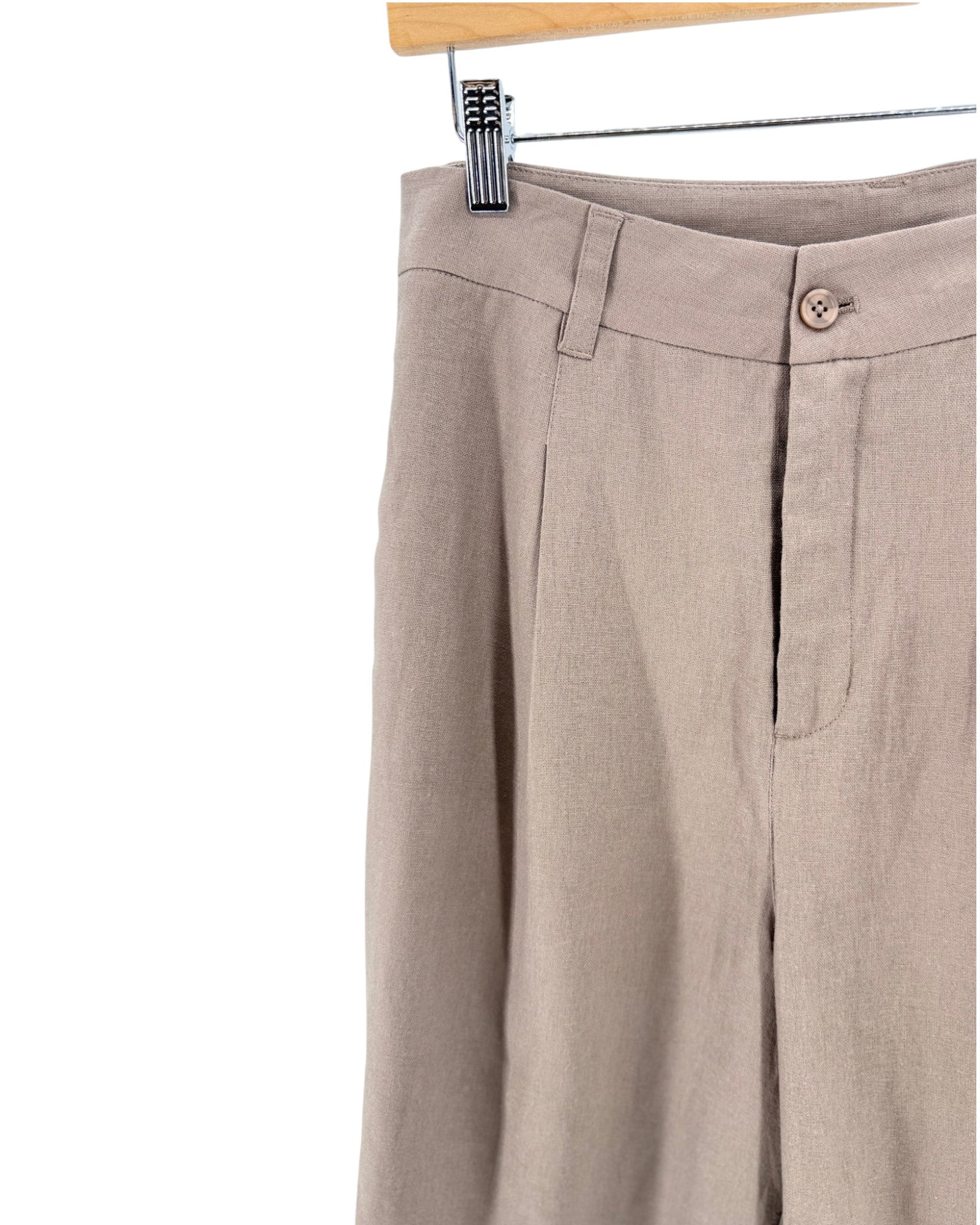 Close-up of beige linen shorts hanging from a wooden hanger with a metal clip, showing the waistband, front fly, and side pocket on a white background.