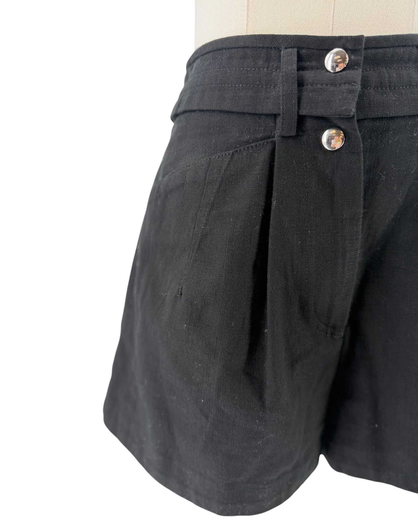 Close-up of black shorts on a mannequin torso, showing the waistband with two silver buttons and pleated front detail against a white background.
