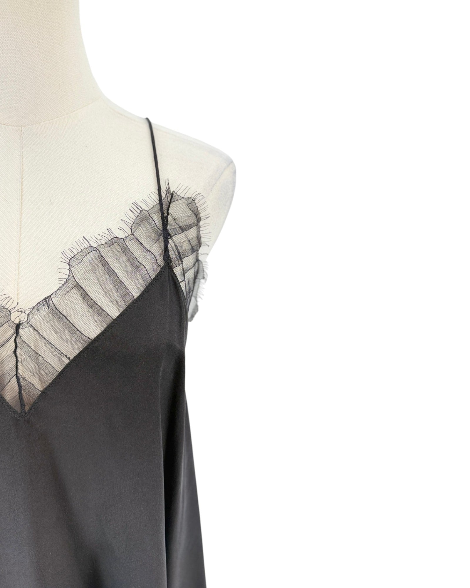 A close-up of a black satin slip dress displayed on a cream mannequin torso against a white background, showing thin straps and a sheer black ruffled lace neckline.