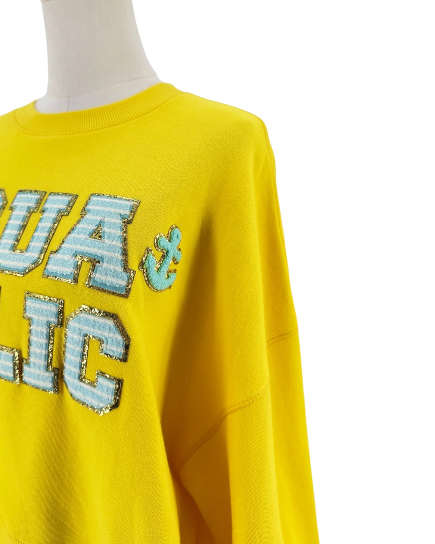 A close-up of a bright yellow crewneck sweatshirt on a mannequin, featuring large sequined letters in light blue and silver on the chest.