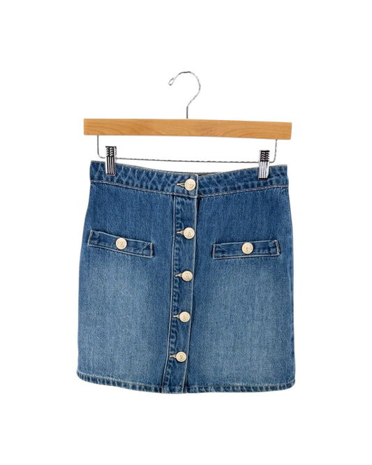 A blue denim mini skirt with a button-down front and two buttoned flap pockets, hanging from a wooden hanger against a plain white background.