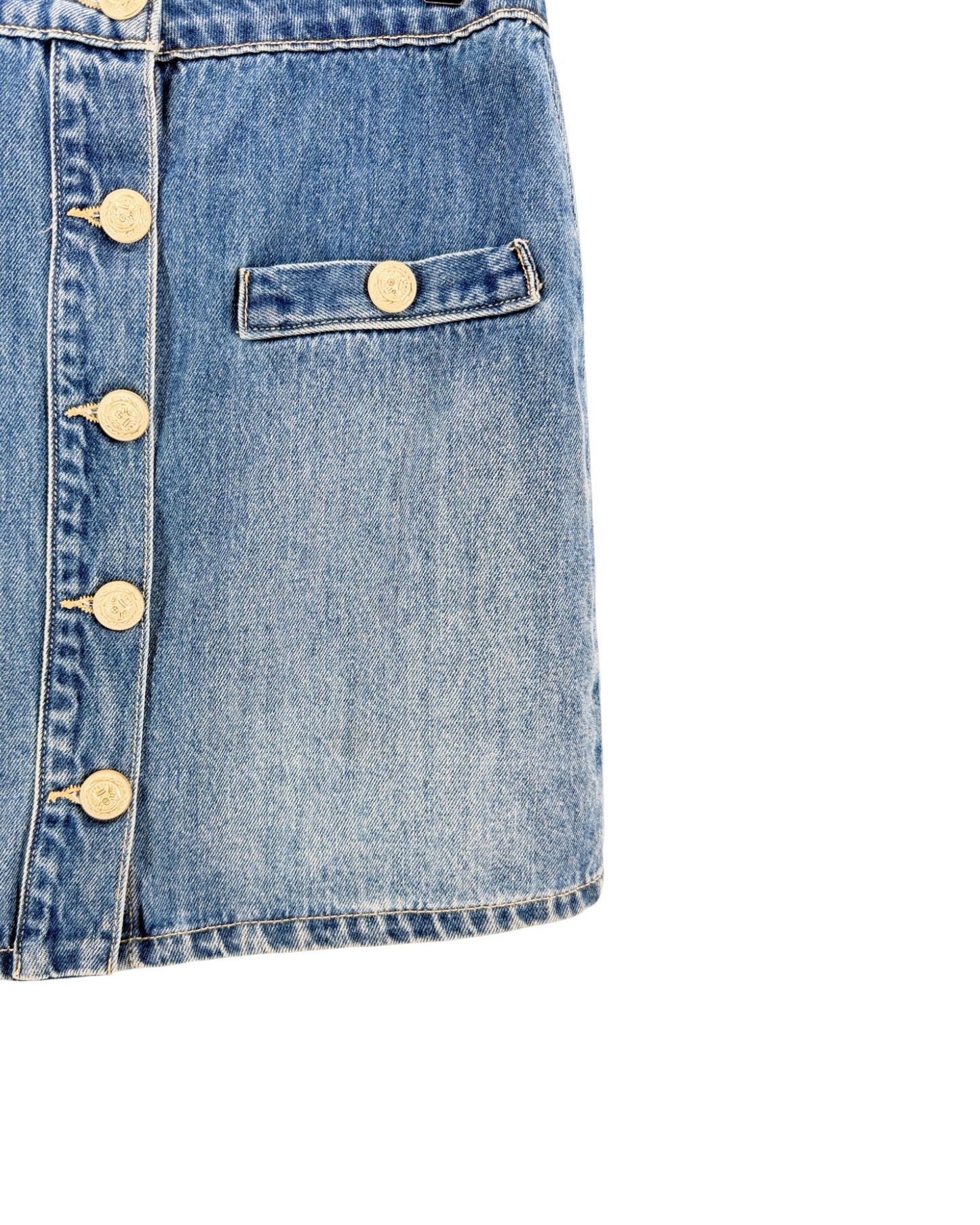 Close-up of a light blue denim skirt showing a buttoned front, a side pocket with a button tab, and gold-tone buttons against a white background.