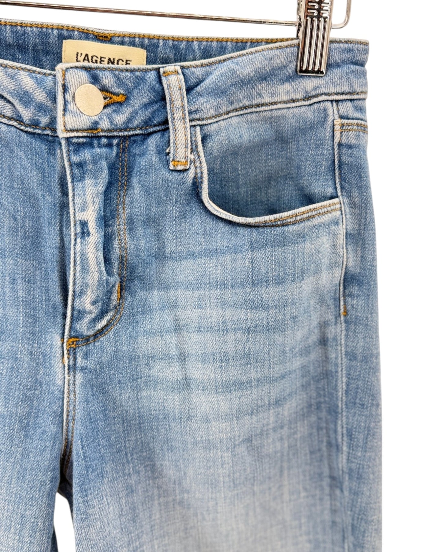 Close-up of light blue denim jeans hanging on a metal hanger, showing the waistband, button, belt loops, front pocket, and a visible L'Agence brand label.
