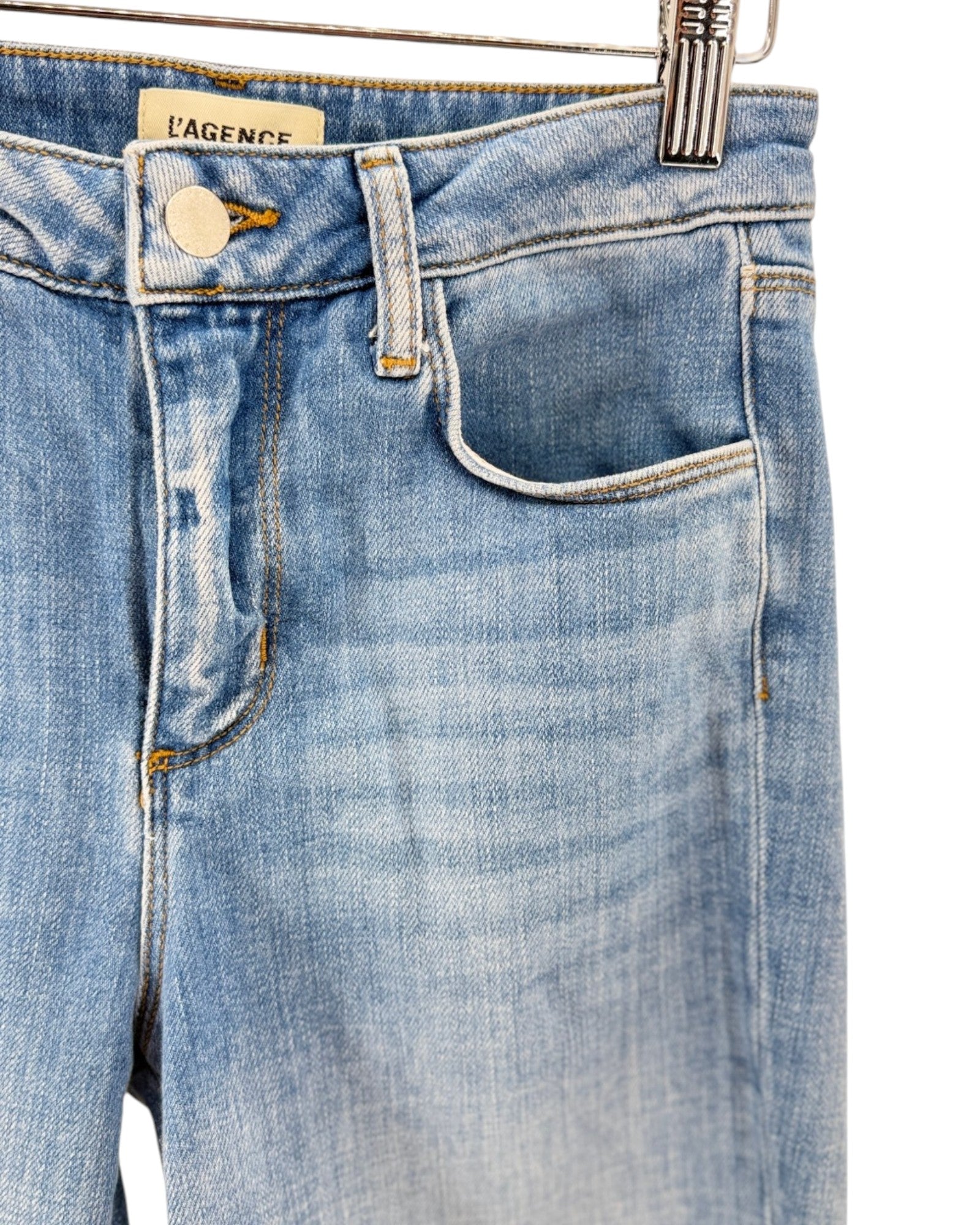 Close-up of light blue denim jeans hanging on a metal hanger, showing the waistband, button, belt loops, front pocket, and a visible L'Agence brand label.