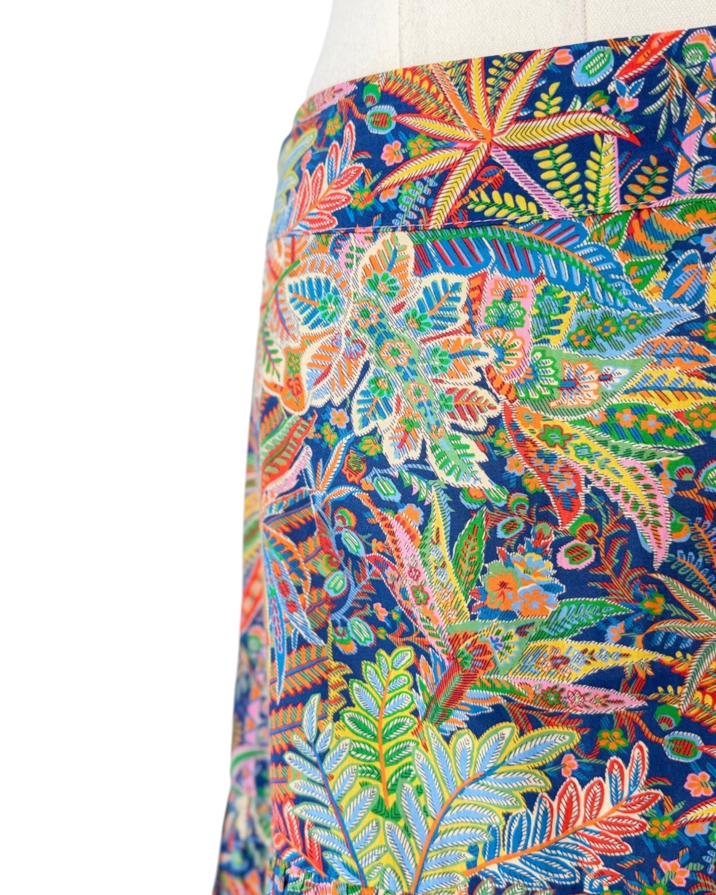 A close-up of a skirt or fabric panel featuring a dense, colorful botanical and floral print on a dark navy background. The pattern includes green, orange, yellow, pink, and blue leaves and flowers, displayed on a mannequin torso against a white background.