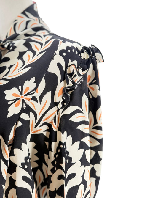 A close-up of a long-sleeve blouse on a mannequin, featuring a black base with a cream and orange floral pattern and ruffled details on the shoulder.