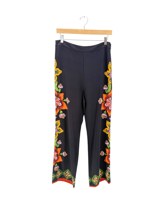 Black wide-leg pants with colorful floral and butterfly patterns down the sides, hanging from a wooden hanger against a white background.
