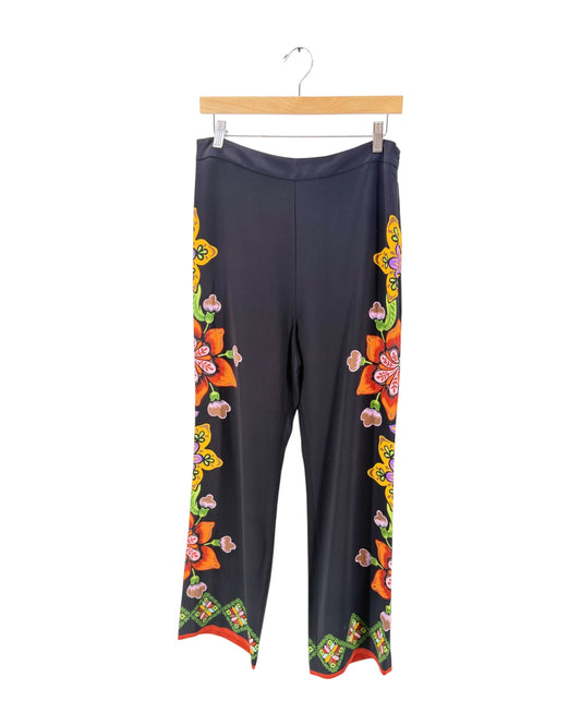 Black wide-leg pants with colorful floral and geometric patterns down the sides and hem, hanging on a wooden hanger against a white background.