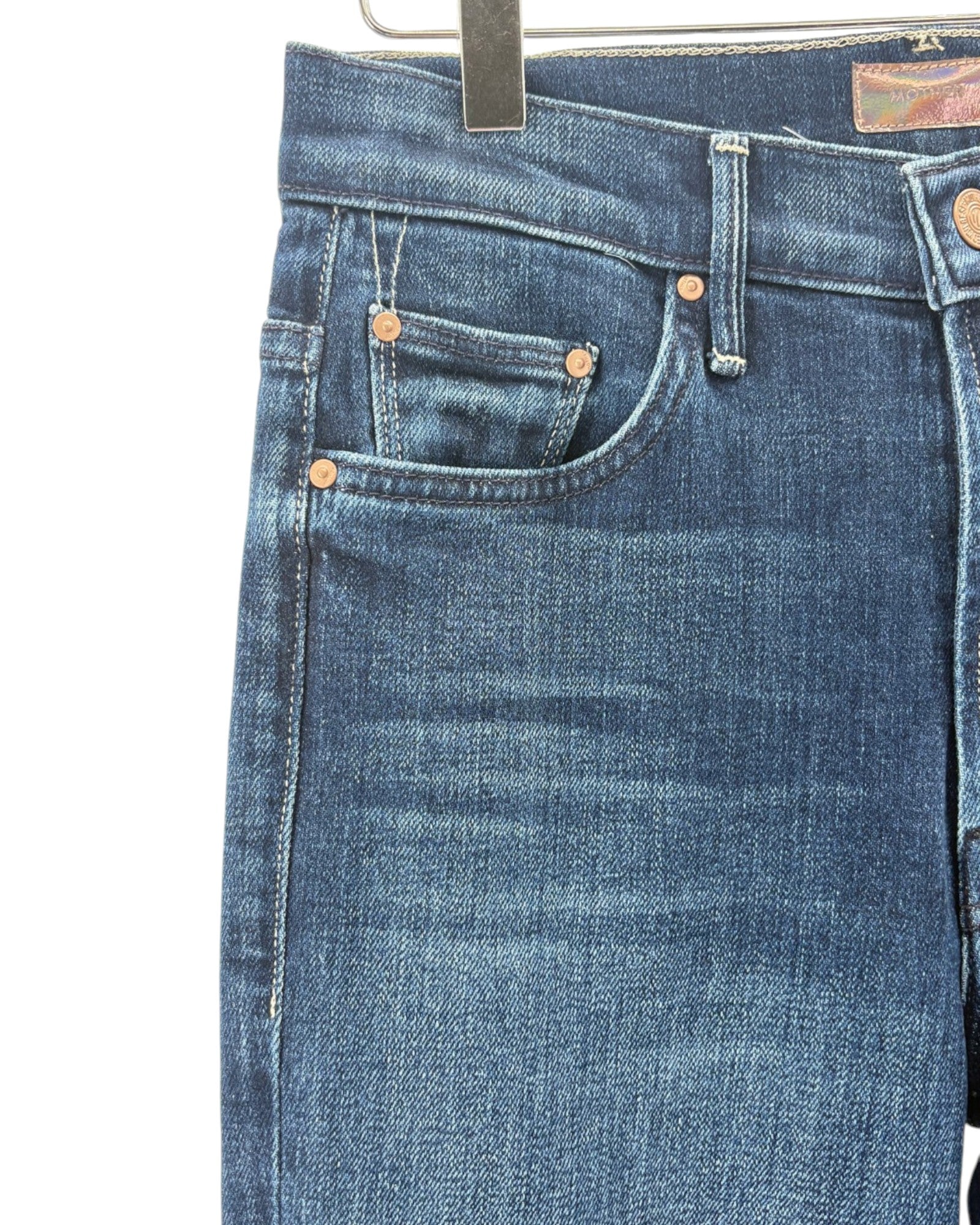 Close-up of a pair of blue denim jeans hanging on a black hanger, showing the front pocket, stitching, rivets, and waistband details against a plain white background.