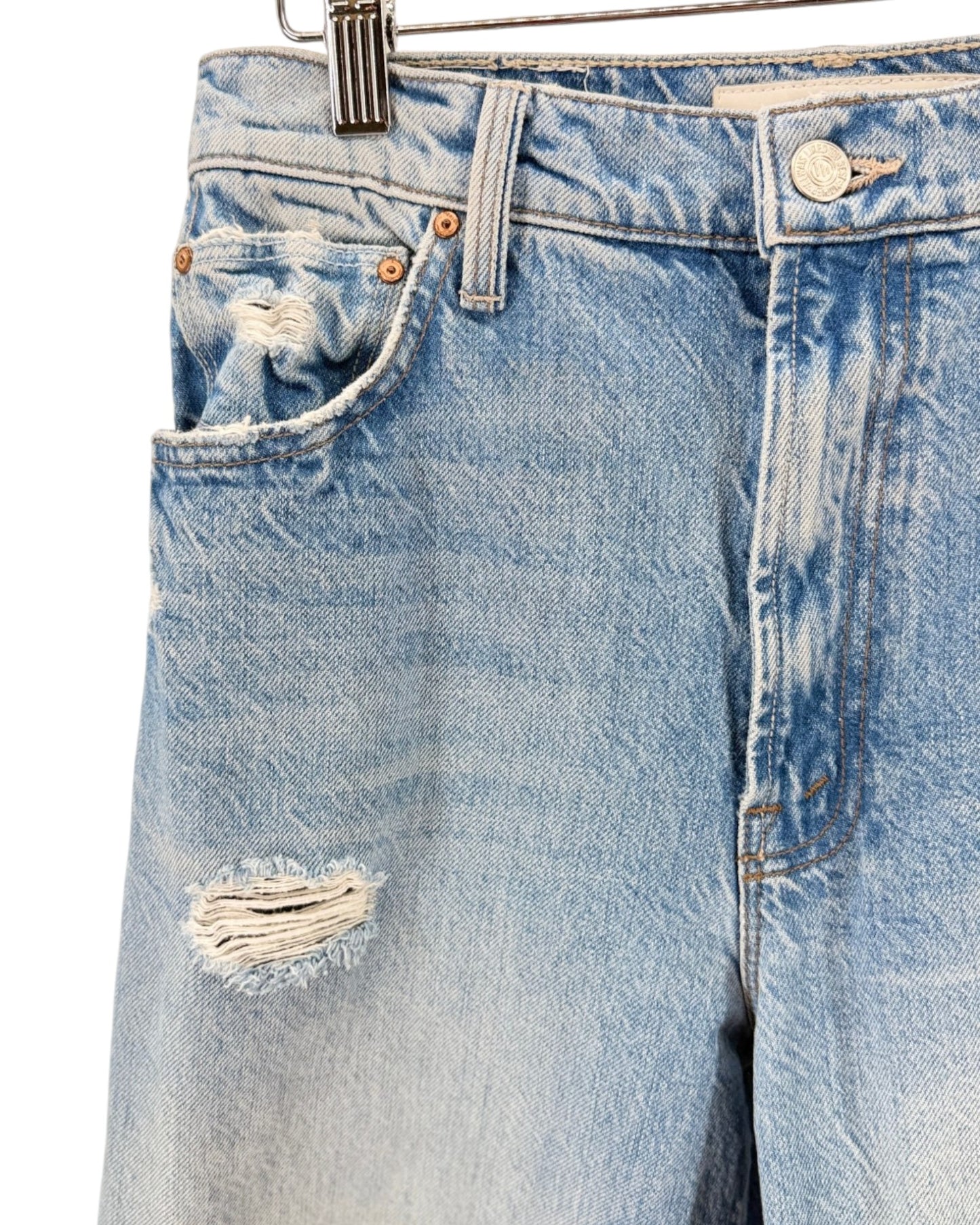 Close-up of light-wash distressed denim shorts showing the front pocket, button, rivets, and a frayed rip on the leg, hanging against a white background.