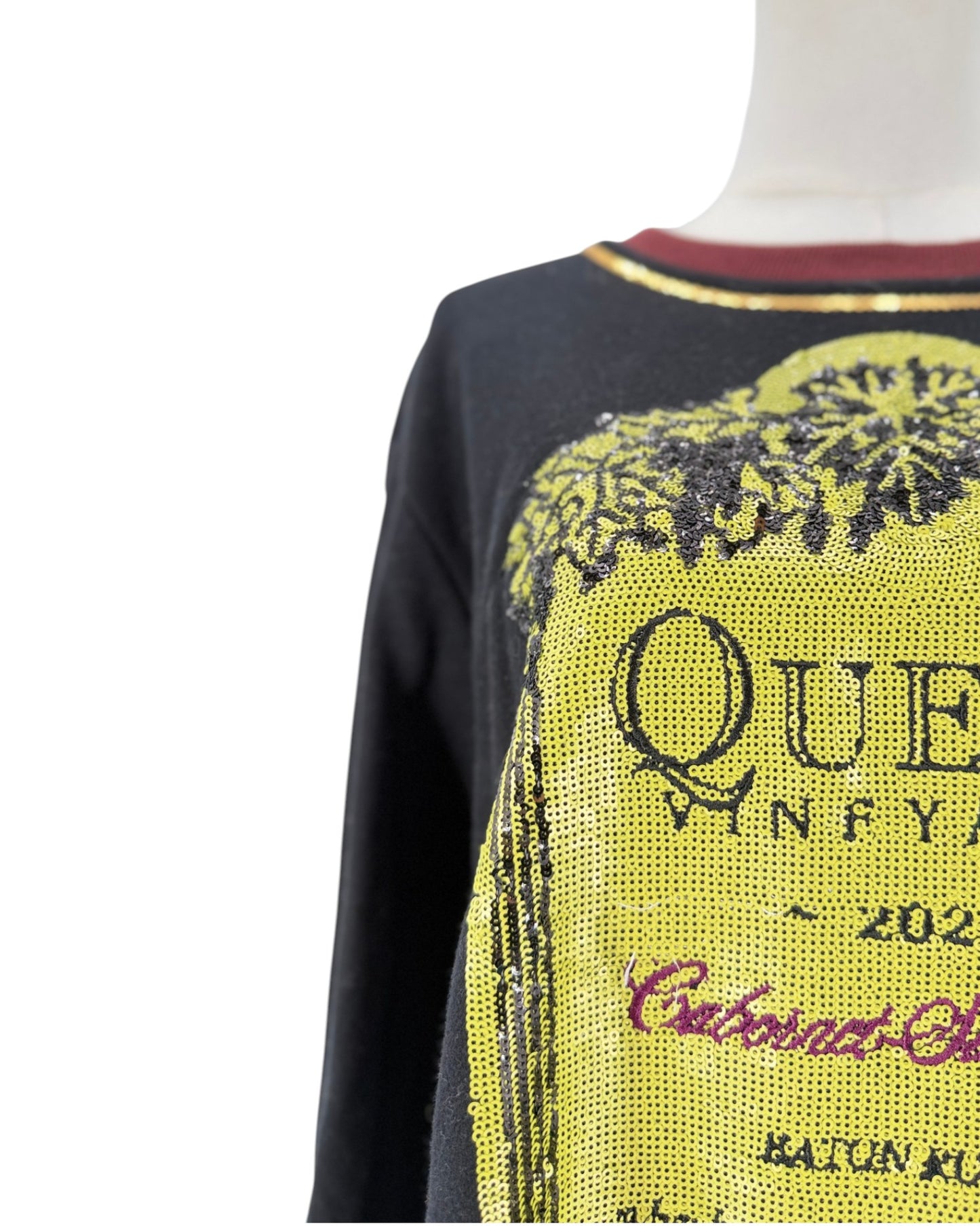 A close-up of a black long-sleeve top on a mannequin torso, featuring a large yellow sequined graphic with black text and a red collar trim.