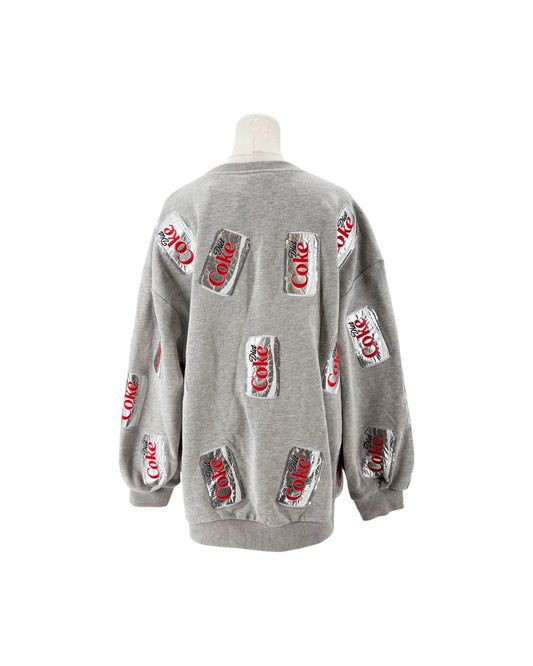A light gray crewneck sweatshirt displayed on a mannequin torso, featuring multiple printed Coca-Cola can graphics on the front and sleeves.