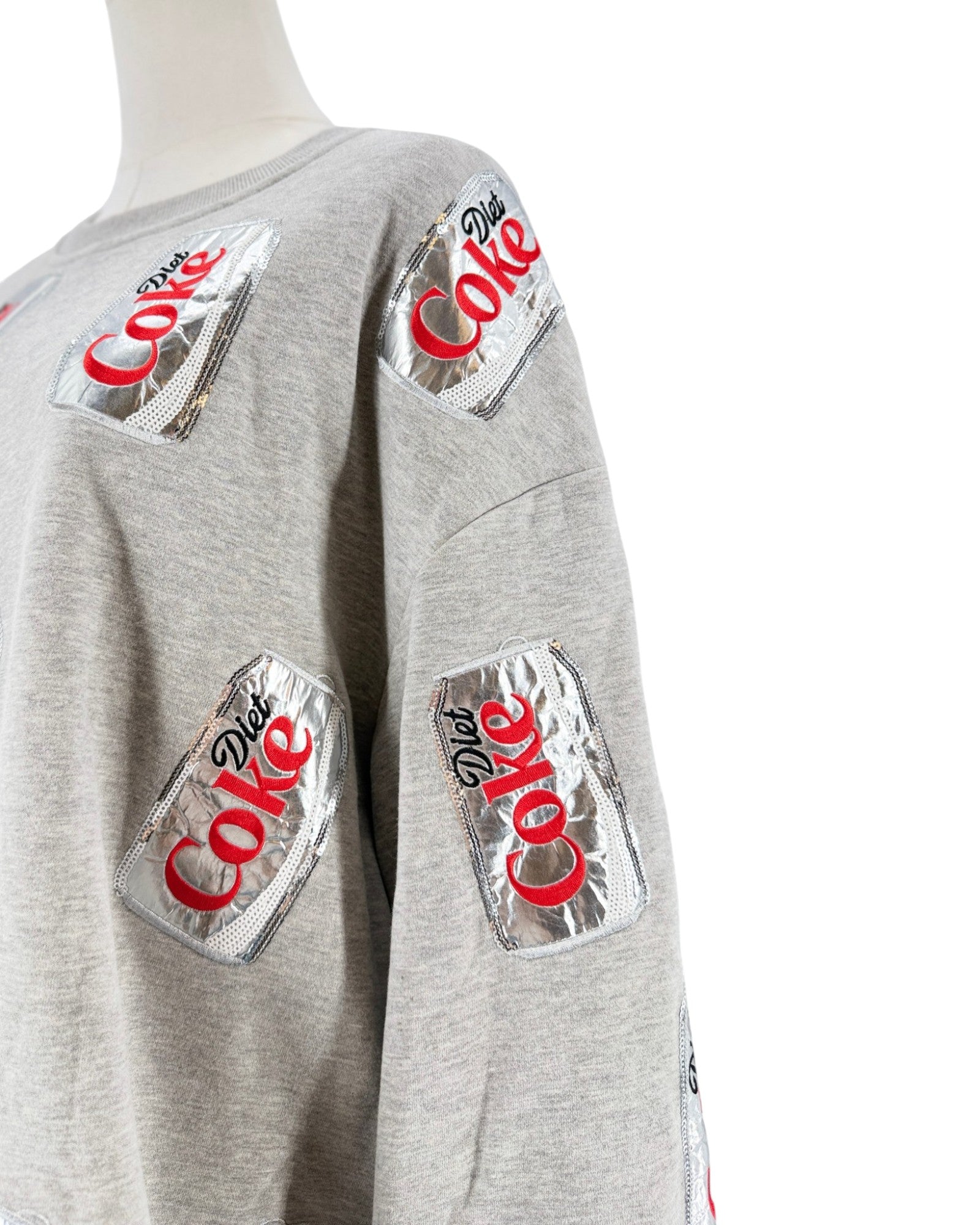 A close-up of a light gray heather sweatshirt on a mannequin, featuring multiple metallic silver patches with the red and black Diet Coke logo printed on the chest and sleeves.